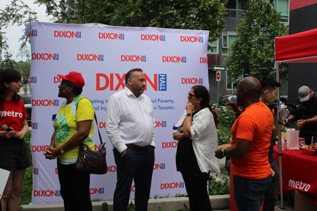 a-group-of-people-stand-and-converse-in-front-of-a-dixon-hall-backdrop-at-an-outdoor-event-some-are-wearing-branded-shirts-and-a-red-tent-with-refreshments-is-visible-in-the-background-