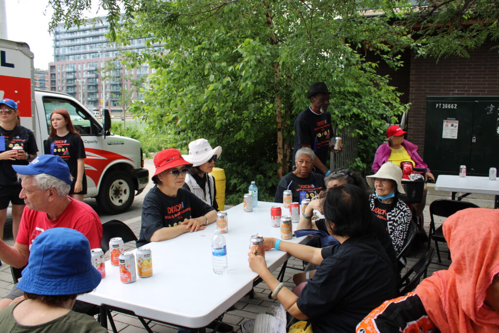 a-group-of-people-wearing-dixon-walk-roll-challenge-2024-t-shirts-sit-at-outdoor-tables-drinking-canned-beverages-and-chatting-a-delivery-truck-and-modern-buildings-are-visible-in-the-background-