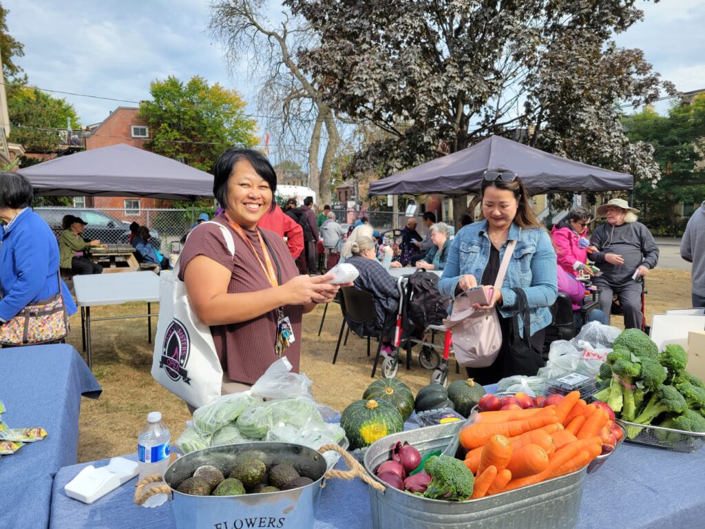 Two women smiling and shopping for fresh vegetables at an outdoor farmers market, with tables of produce, tents, and people socializing in the background.