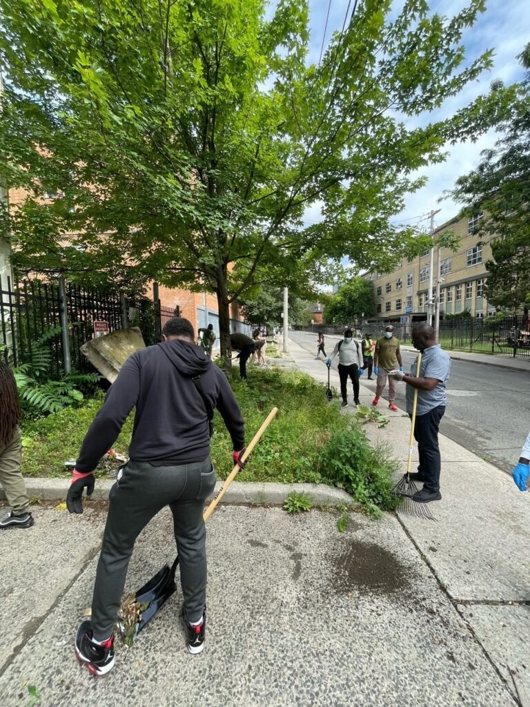 a-group-of-people-is-participating-in-a-community-cleanup-using-shovels-and-rakes-to-clear-overgrown-weeds-and-debris-from-a-sidewalk-area-trees-and-a-school-building-are-visible-in-the-background-