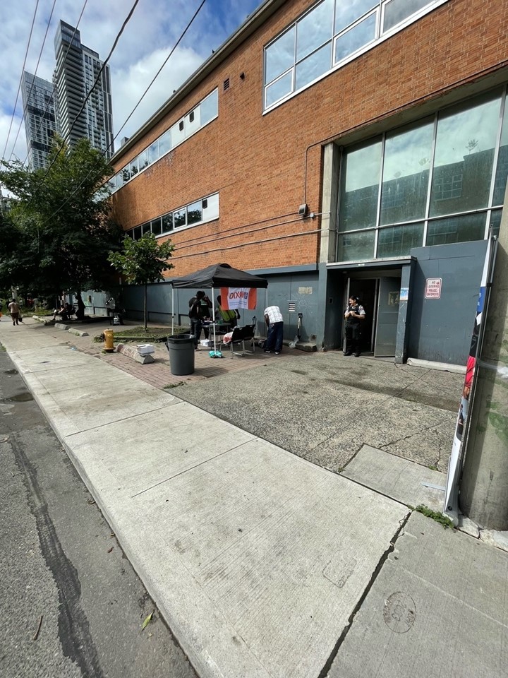 a-brick-building-with-large-windows-and-an-open-entrance-is-visible-outside-a-small-tent-with-a-dixon-hall-banner-is-set-up-where-a-few-people-are-gathered-a-security-officer-stands-near-the-doorway-and-a-trash-bin-is-placed-on-the-sidewalk-tall-buildings-are-seen-in-the-background