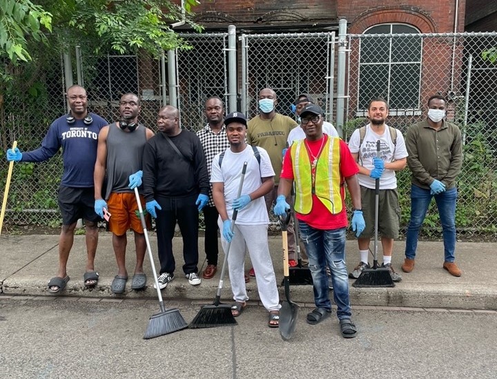 a-group-of-volunteers-is-posing-for-a-photo-on-a-sidewalk-holding-brooms-and-wearing-gloves-some-are-wearing-safety-vests-and-face-masks-a-chain-link-fence-and-an-old-brick-building-with-arched-windows-are-visible-in-the-background