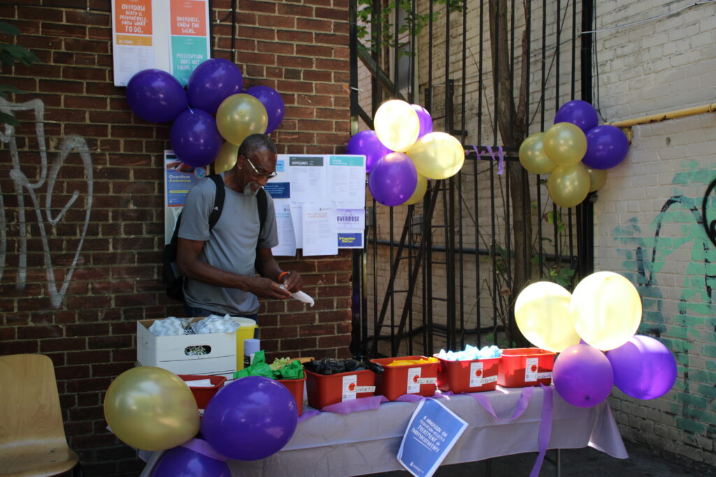 a-man-with-a-backpack-is-standing-at-an-outdoor-table-decorated-with-purple-and-gold-balloons-selecting-items-from-labeled-bins-the-table-holds-harm-reduction-supplies-and-a-brick-wall-behind-him-is-covered-with-informational-posters-about-overdose-awareness