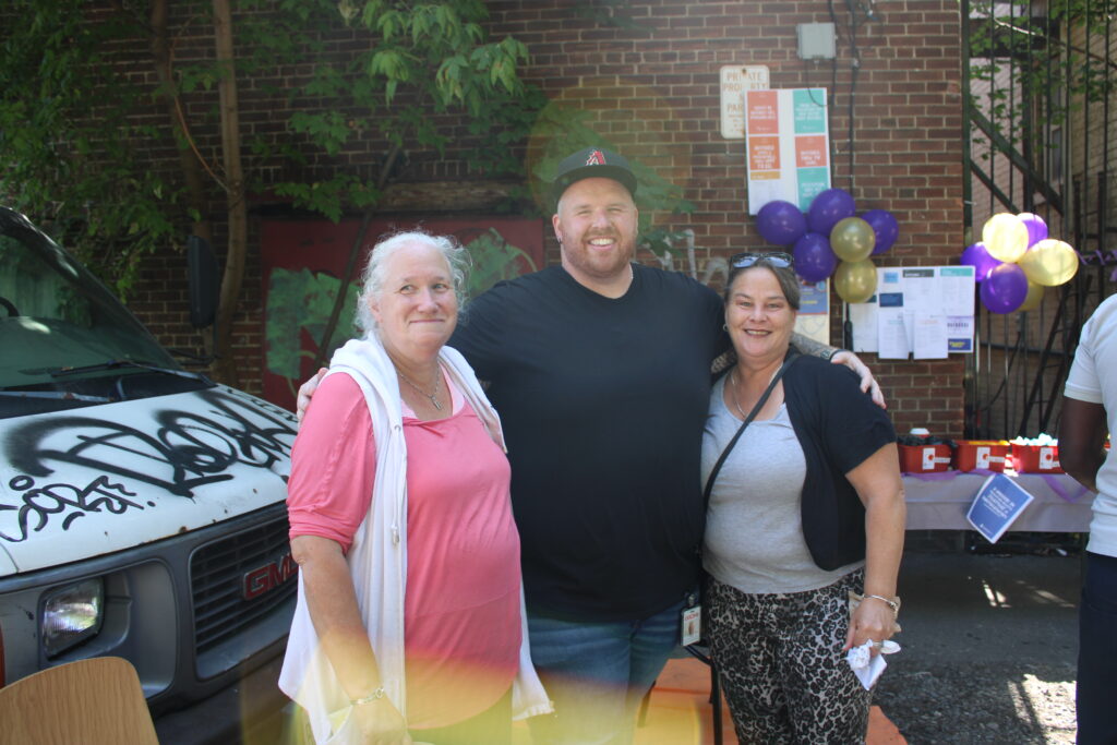 three-people-are-standing-together-outdoors-smiling-at-the-camera-behind-them-a-table-decorated-with-purple-and-gold-balloons-holds-supplies-and-a-brick-wall-with-posters-is-visible-a-graffiti-covered-van-is-parked-nearby