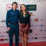 Two event staff members pose together on the red carpet in front of a branded backdrop that reads "Music for Life." They are wearing name tags and professional attire, smiling for the camera. The backdrop features various sponsor logos.