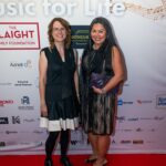 Two women pose on the red carpet at the "Music for Life" event, standing in front of a backdrop featuring sponsor logos and musical notes.