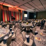 A banquet hall set up for an event, featuring round tables with elegant table settings, gift bags, and floral centerpieces. A stage with a piano and podium is illuminated by warm lighting in the background.