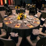 A round banquet table elegantly set with silverware, wine glasses, and folded napkins. A floral centerpiece with yellow flowers and candles sits in the middle. Gift bags with tissue paper are placed on the chairs, and event programs labeled "Music for Life" rest on the table. The stage with warm lighting is visible in the background.