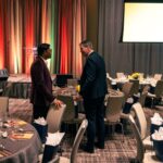 Two men in formal attire engaged in conversation near a table at a decorated banquet hall, with a stage and a presentation screen in the background.