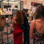 Guests browse a silent auction display featuring gift baskets, framed pictures, and various items wrapped in cellophane. The display is illuminated by small candles, and attendees in elegant attire examine the auction details.