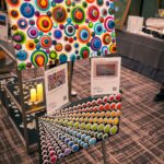 Two colorful abstract paintings featuring vibrant circular patterns are displayed on a stand at an event. Informational cards with details about the artwork are placed below each painting. The background shows a venue with decorative elements and auction items.