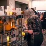 A woman in an elegant black lace dress holds a wine glass while browsing items displayed on shelves at a formal event. The shelves contain various gift baskets, candles, and decorative items. The background shows other guests mingling.