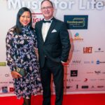 A well-dressed couple poses on the red carpet at the "Music for Life" event, standing in front of a backdrop featuring sponsor logos and event branding.