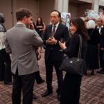 A group of elegantly dressed people engaged in conversation at a formal event, holding drinks in a well-lit venue with decorative displays in the background.