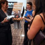 A woman in a dark suit serves a tray of small appetizers to a group of elegantly dressed women holding wine glasses. A sign in the background promotes a music school with an image of two musicians playing guitars.