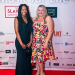 Two women posing and smiling on the red carpet at the "Music for Life" event, with a branded backdrop featuring sponsor logos behind them. One woman is wearing a black dress and holding a black purse, while the other is in a colorful floral dress with teal heels.