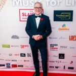 A man in a navy tuxedo with a bow tie and red poppy pin poses on the red carpet at the "Music for Life" event, standing in front of a branded backdrop featuring sponsor logos.