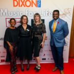 Four elegantly dressed individuals pose on the red carpet in front of a "Music for Life" event backdrop featuring sponsor logos. The group includes three women in stylish black outfits and a man in a blue suit with an orange tie, all smiling for the camera.
