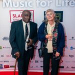 An older man and woman pose together on the red carpet at a "Music for Life" event, holding glasses of wine. The man wears a suit and tie while holding a cane, and the woman wears a patterned scarf and cardigan. The event backdrop features sponsor logos.