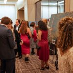 Guests at a formal event mingle and admire artwork on display. Several people, dressed in elegant attire, engage in conversation while others observe paintings set on easels. The setting is a well-lit indoor venue with a sophisticated atmosphere.