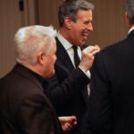 A group of well-dressed men in suits enjoying appetizers at a formal event, with one man laughing while taking a bite. The atmosphere appears lively and social.