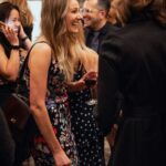 A woman in a floral dress laughs while holding a glass of red wine at a formal event. Other guests in elegant attire are engaged in conversation in the background, creating a lively and social atmosphere.