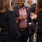 A man in a checkered blazer with a red poppy pin is smiling and holding a wine glass while engaged in conversation with a group of people at a formal event. Other attendees in elegant attire are mingling in the background.