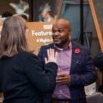 A man in a checkered blazer with a red poppy pin holding a wine glass, engaged in conversation with a woman at a formal event, with a promotional sign in the background.