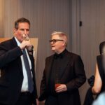 Two men in formal attire engaged in conversation at a networking event. One man in a dark suit sips from a wine glass, while the other in a black tuxedo with a bow tie holds a cocktail. A colorful painting is visible in the background.