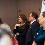 A woman in a black dress and a man in a suit stand together at a formal event, engaged in conversation and smiling, with other attendees visible in the background.