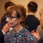 A woman wearing glasses and a floral blouse is engaged in conversation at a formal networking event, with other attendees mingling in the background.