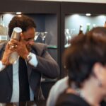 A bartender in a suit energetically shaking a cocktail shaker behind a bar, with shelves of glassware and bottles in the background. A blurred crowd is visible in the foreground.