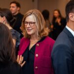 A woman wearing glasses and a magenta jacket is smiling while engaged in conversation at a social event. Other attendees are mingling in the background.