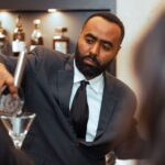 A bartender in a suit and tie carefully pouring a drink into a martini glass at a formal event, with liquor bottles displayed in the background.