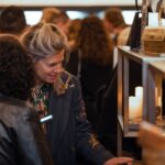 A woman in a dark floral-patterned blazer browses items on a shelf at a crowded event, appearing engaged in conversation with another attendee.