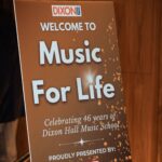 A promotional sign for the "Music For Life" event, celebrating 46 years of Dixon Hall Music School. The sign features a warm golden design with sparkling details and includes the event's title, a welcome message, and sponsorship information.