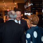 A man in a suit is engaged in conversation with two other attendees at a formal event, with a warmly lit venue in the background.