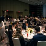 Guests seated at elegantly set tables, engaged in conversation at a formal banquet event. The room is dimly lit with warm lighting, and floral centerpieces with candles decorate the tables. Attendees are dressed in formal attire, some reading event programs while others are mingling.