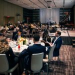 Guests seated at a round table in a banquet hall, engaged in conversation and reading event programs. The stage in the background is set with microphones and a piano, with elegant lighting and decorations.