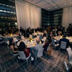 A banquet hall filled with elegantly set tables and guests dressed in formal attire. The room features floor-to-ceiling windows and decorative hanging lights, creating a sophisticated atmosphere. Attendees are engaged in conversation while enjoying their meals.