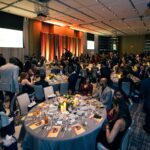 A large banquet hall filled with elegantly dressed guests seated at round tables adorned with candles and floral centerpieces. The stage in the background features a grand piano and a backdrop with warm lighting, while attendees engage in conversation and enjoy the event.
