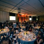 A large banquet hall filled with elegantly decorated tables and guests socializing, with a stage, a grand piano, and presentation screens in the background.