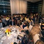 A large banquet hall filled with elegantly dressed guests seated at round tables adorned with floral centerpieces, candles, and dinnerware. Attendees are engaged in conversation and dining, with a softly lit stage and decorative drapery in the background.
