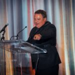 A man in a black suit speaking at a clear podium with microphones, gesturing towards the audience at a formal event. The background features draped curtains.