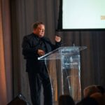 A man dressed in black is speaking at a clear podium with a microphone at the Four Seasons Hotel Toronto. A screen displaying event details is visible in the background, and audience members are seated in the foreground.