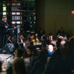 A speaker at a podium addressing a seated audience at a formal dinner event. The dimly lit room has elegant table settings, floral centerpieces, and guests attentively listening.