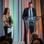 A man in a suit speaks at a podium with the Four Seasons Hotel Toronto logo, while a woman in a beige blazer stands beside him holding papers, smiling. The background features colorful drapery and an audience watching the presentation.