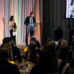 A man in a suit speaks at a podium with a "Four Seasons" logo, while a woman stands beside him holding papers. The audience is seated at elegantly set tables, and a screen displays a live feed of the event.