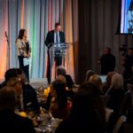 A man in a suit speaks at a podium with a Four Seasons logo, while a woman stands beside him holding papers. The audience is seated at round tables with candles, attentively listening. A camera operator and security personnel are visible in the background.