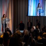 A woman speaks at a podium with a Four Seasons logo, addressing an audience seated at round tables. A large screen behind her displays a live feed of her speech. Security personnel and a camera operator are visible in the background.